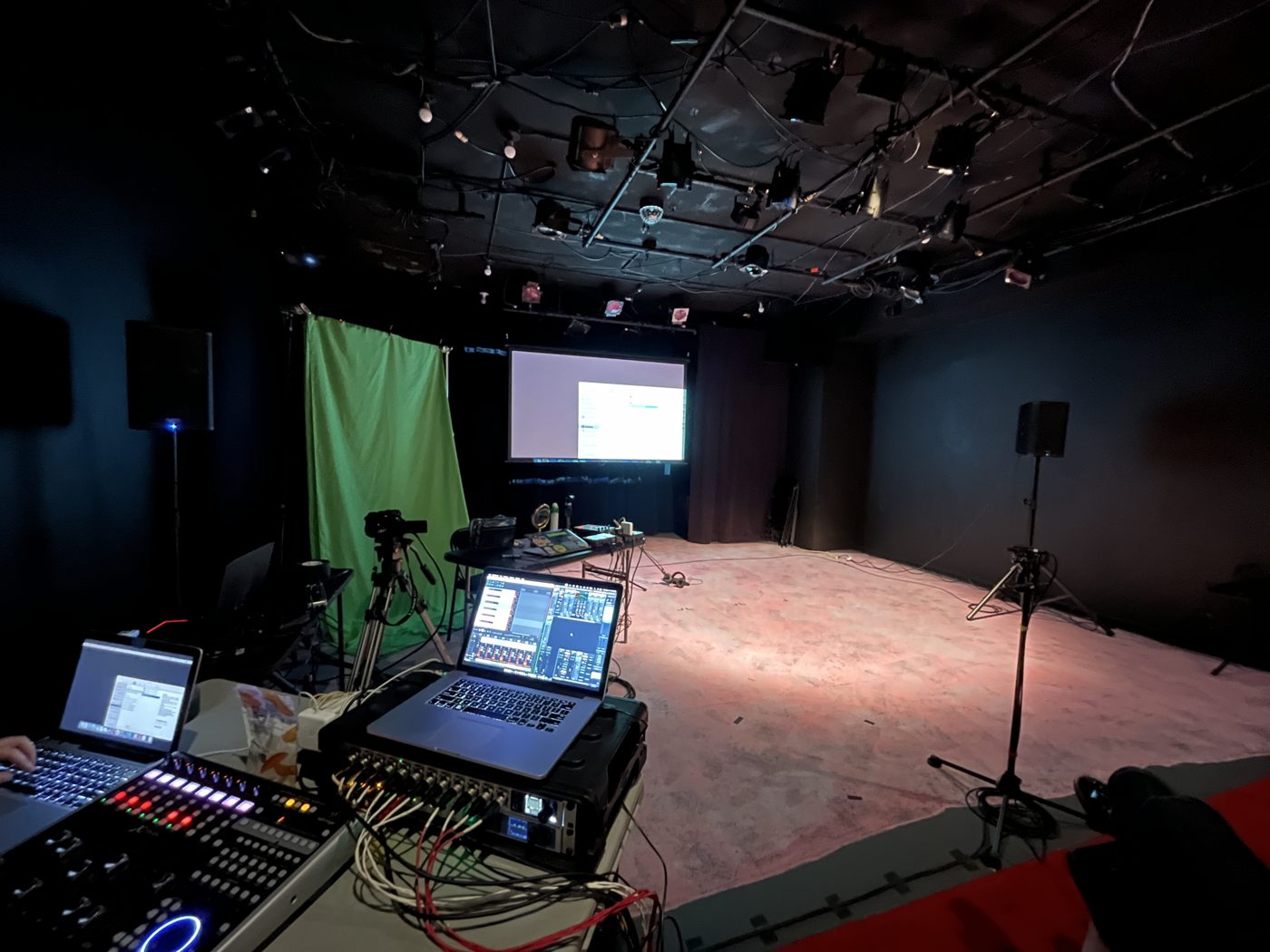 Mixing console and laptops at a front-of-house position facing a black-box stage with projection